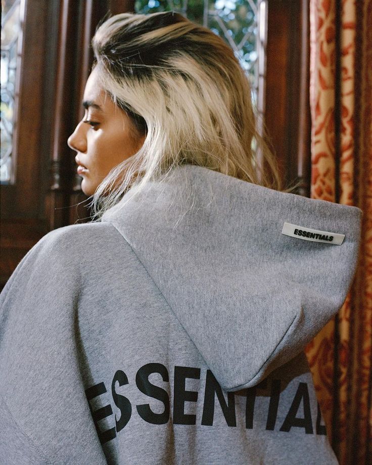Woman wearing a gray hoodie with 'ESSENTIALS' tag, showcasing modern streetwear style against elegant background.