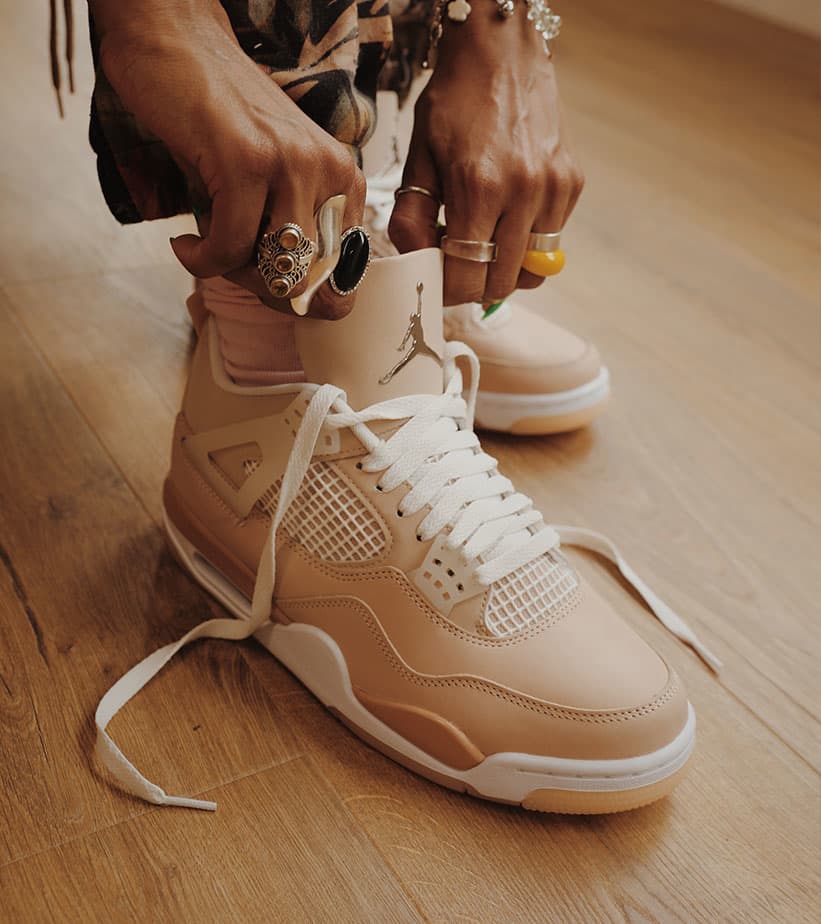 Close-up of hands lacing up beige Air Jordan 4 sneakers on wooden floor, showcasing stylish footwear.