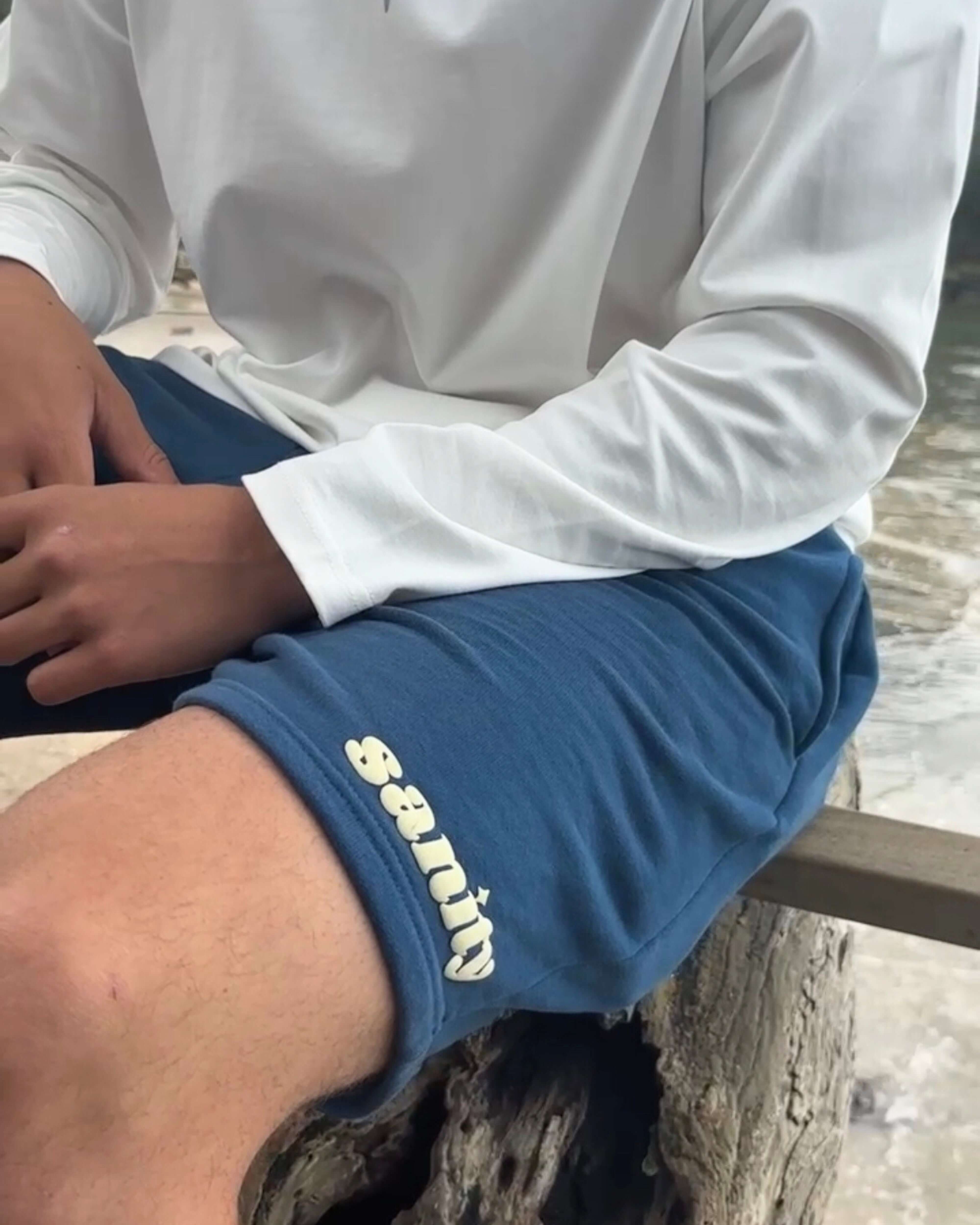 Model wearing Sanity Deep Sea Men's Shorts in navy with white logo, seated outdoors near the water.