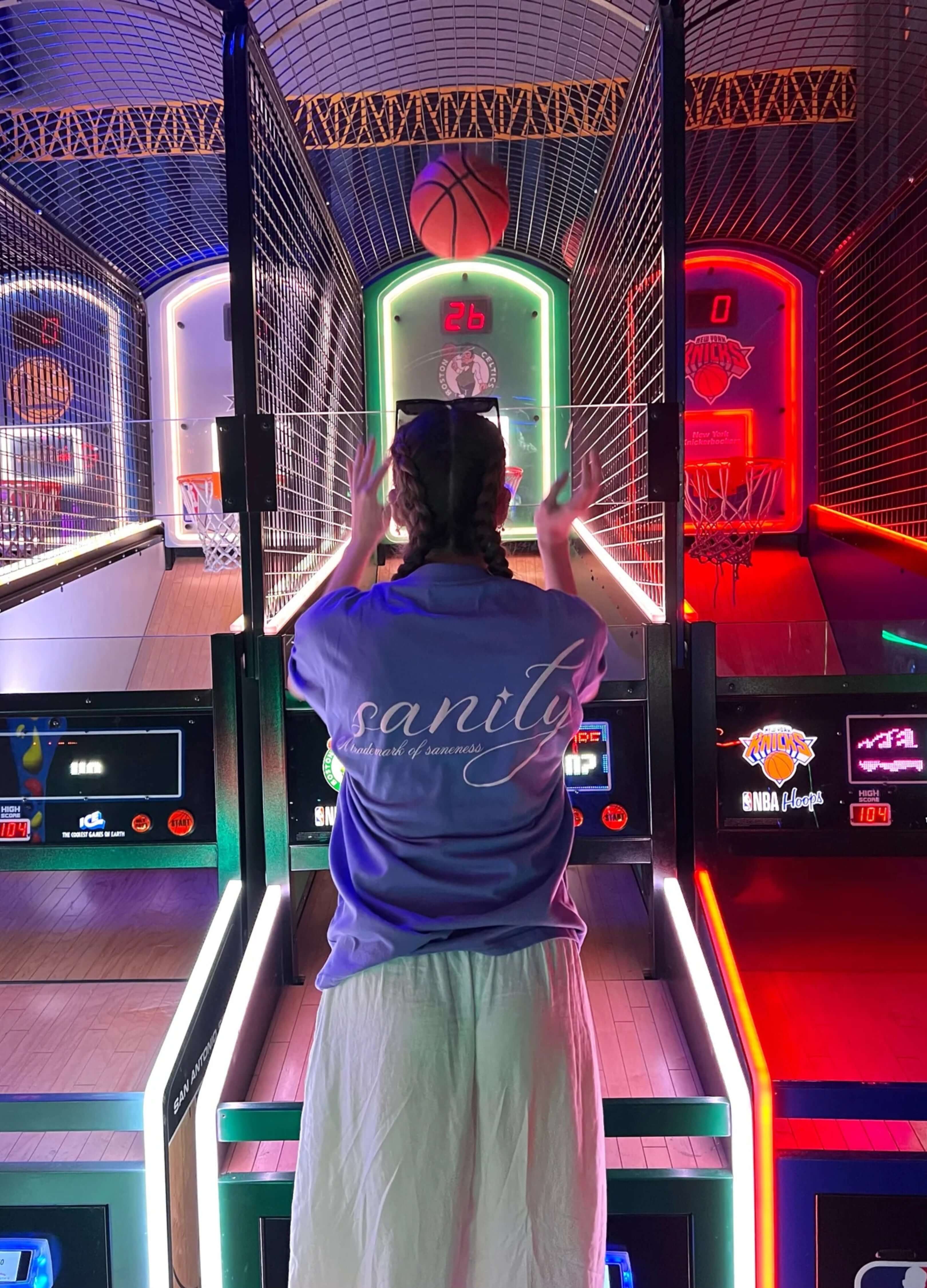 Person wearing a purple tee standing in front of illuminated basketball arcade games, enjoying a vibrant gaming experience.