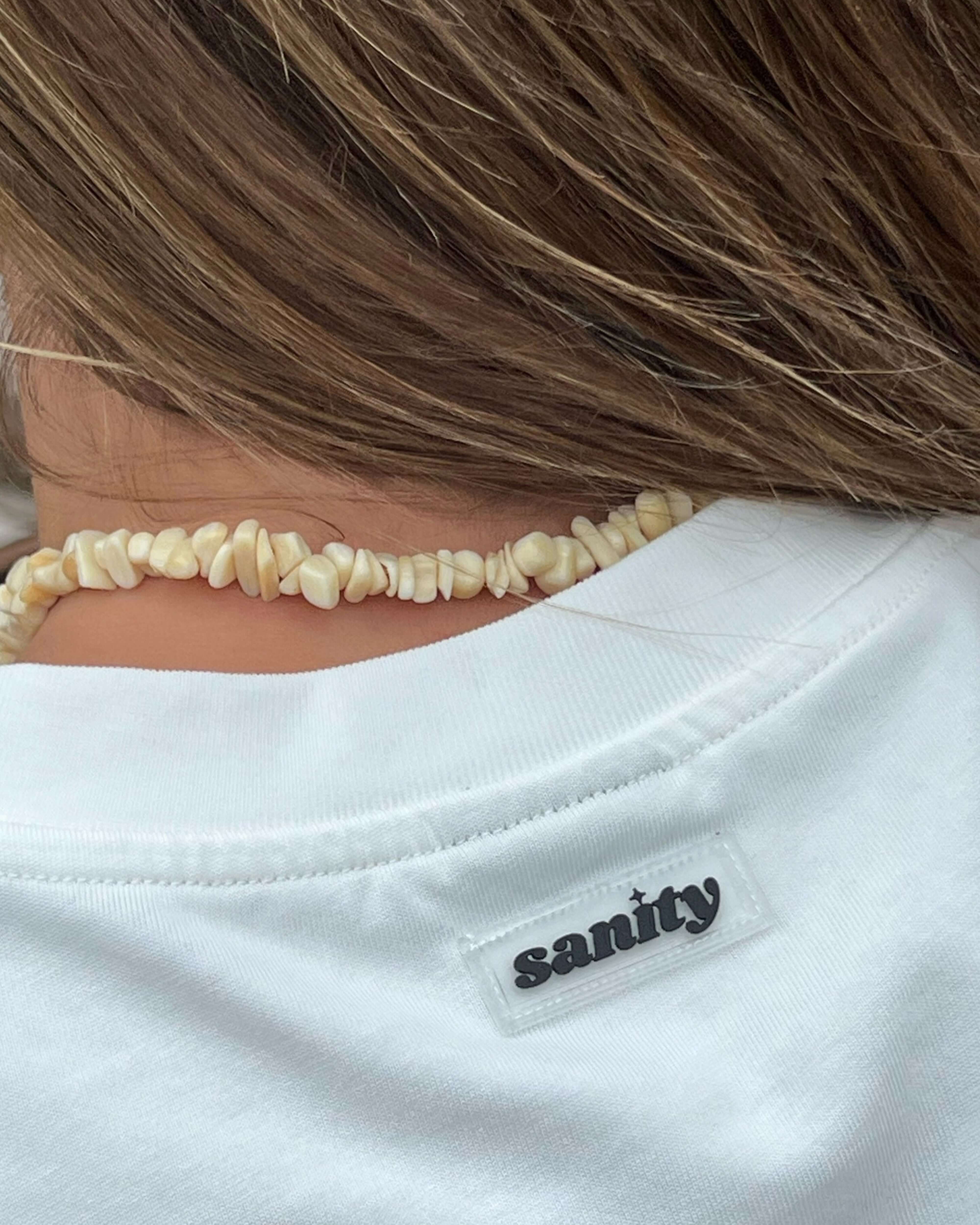 Close-up of the back of a Cloud White Long Sleeve Tee by Sanity, showcasing the logo tag and natural necklace.