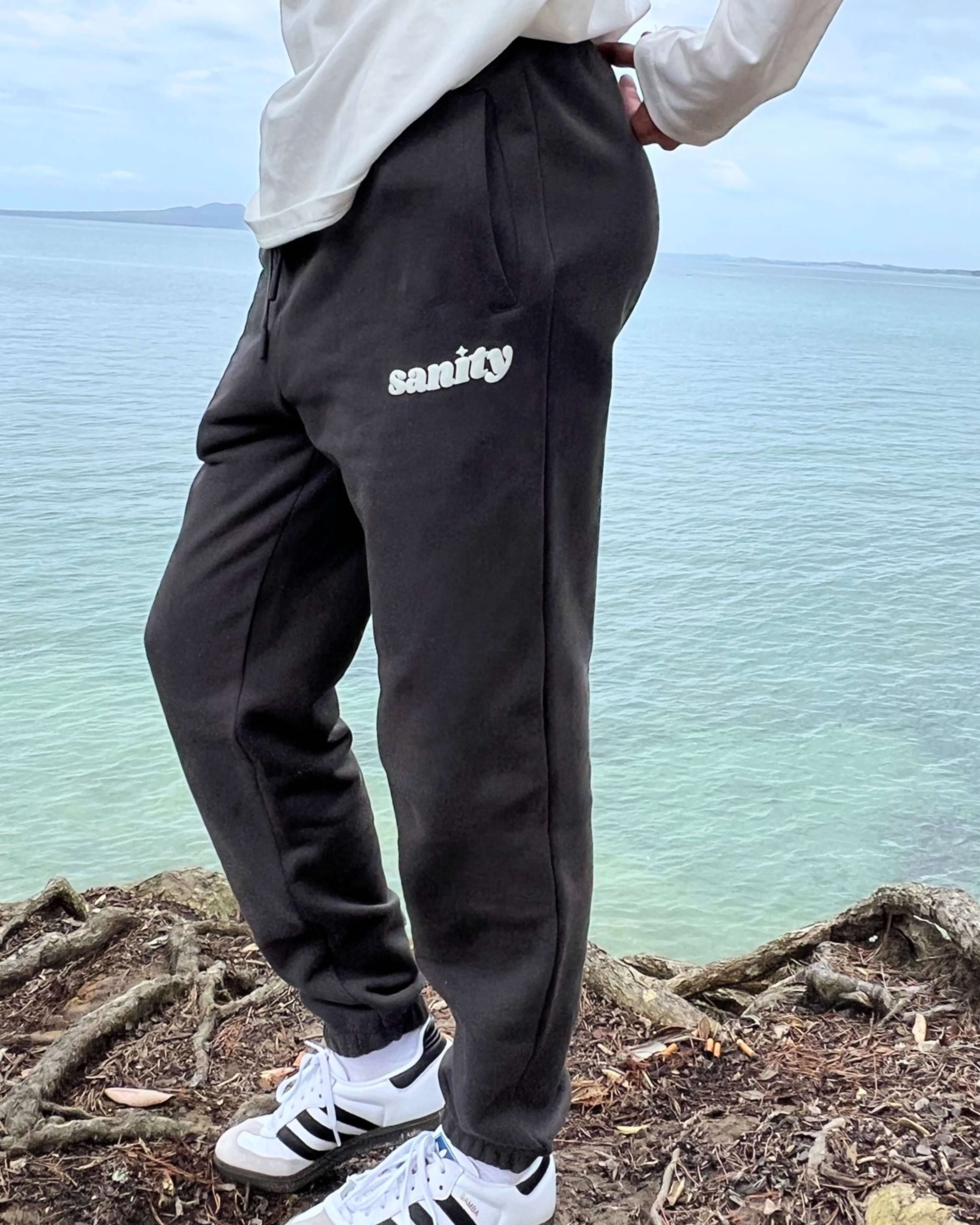 Model wearing Sanity Shadow Trackpants by the water, showcasing sleek design and comfort in deep charcoal.