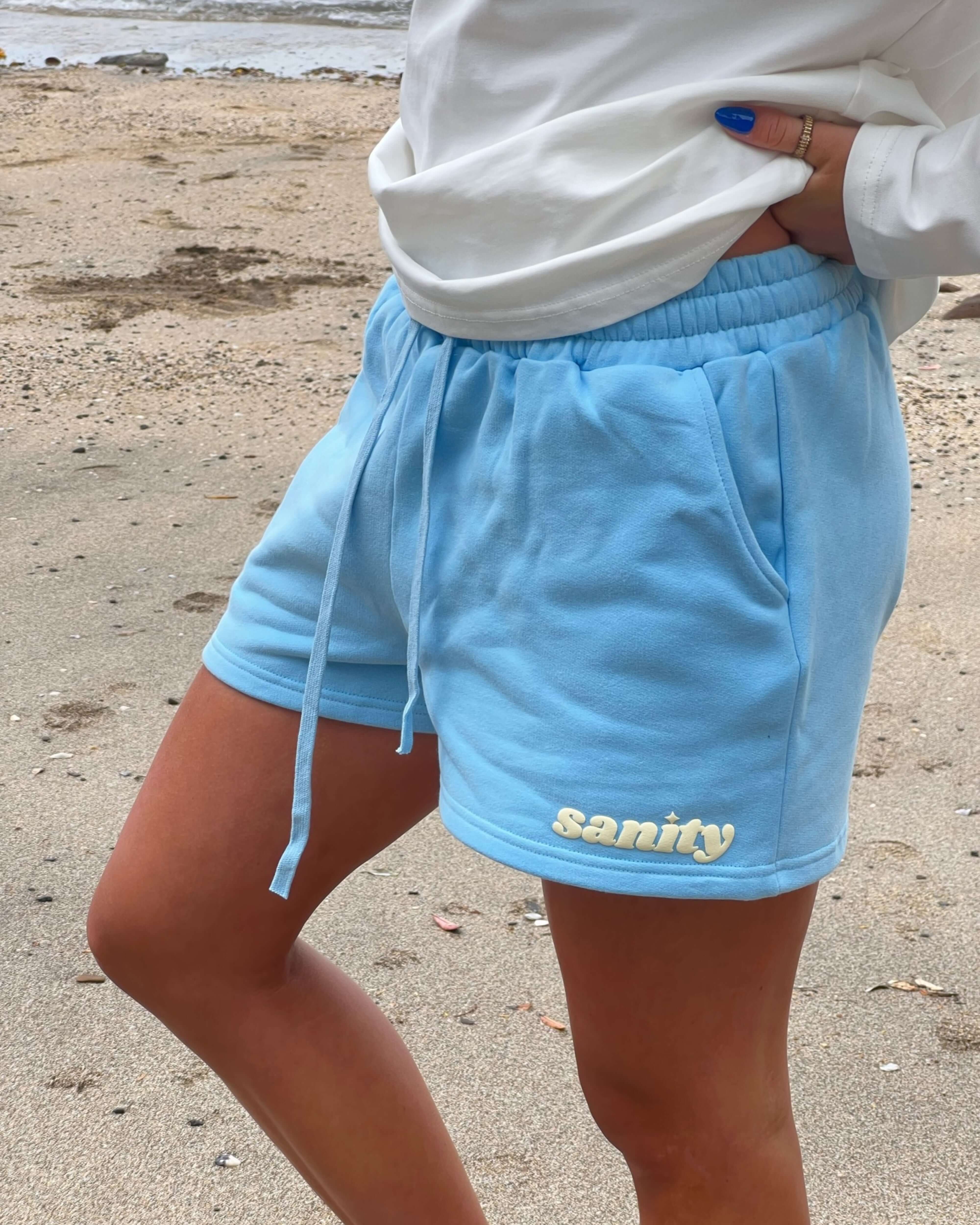 Relaxed high-waisted Ice Blue women's shorts with side pockets and 'sanity' embroidered logo on a beach.