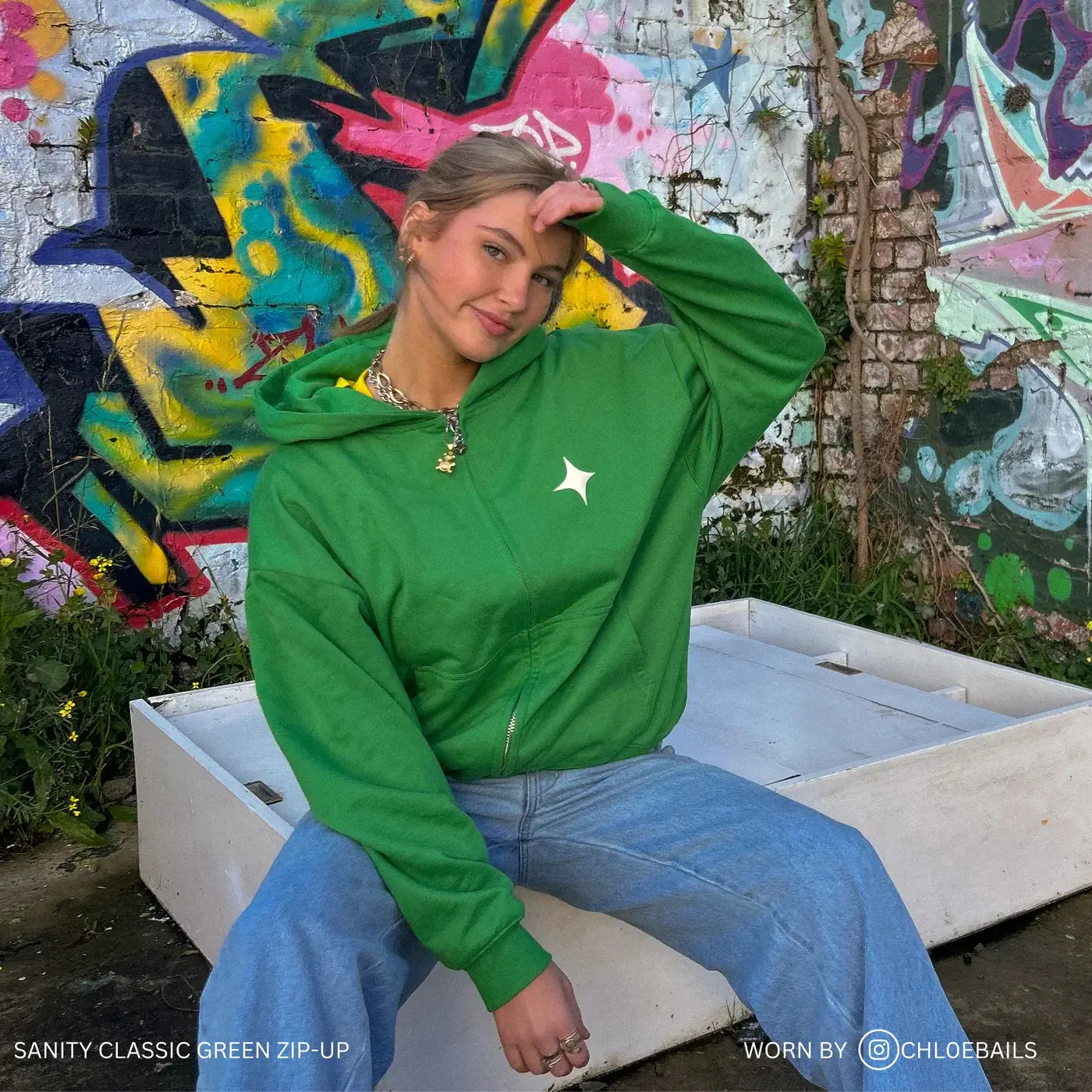 Model wearing a Sanity green zip-up hoodie with white print, showcasing a laid-back style against a colorful graffiti background.