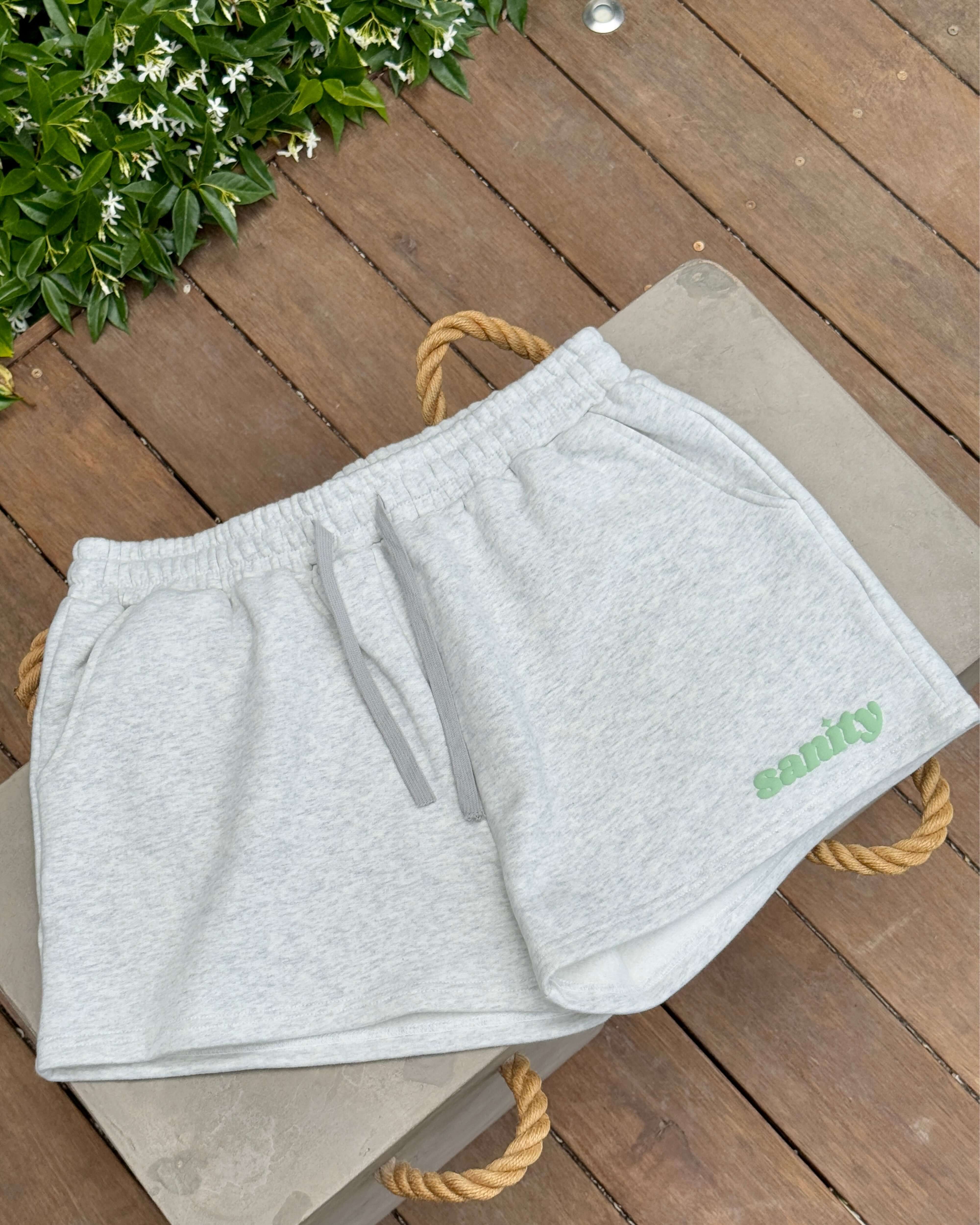 Sanity gray women's shorts with elastic waistband and logo, displayed on a wooden surface surrounded by greenery.