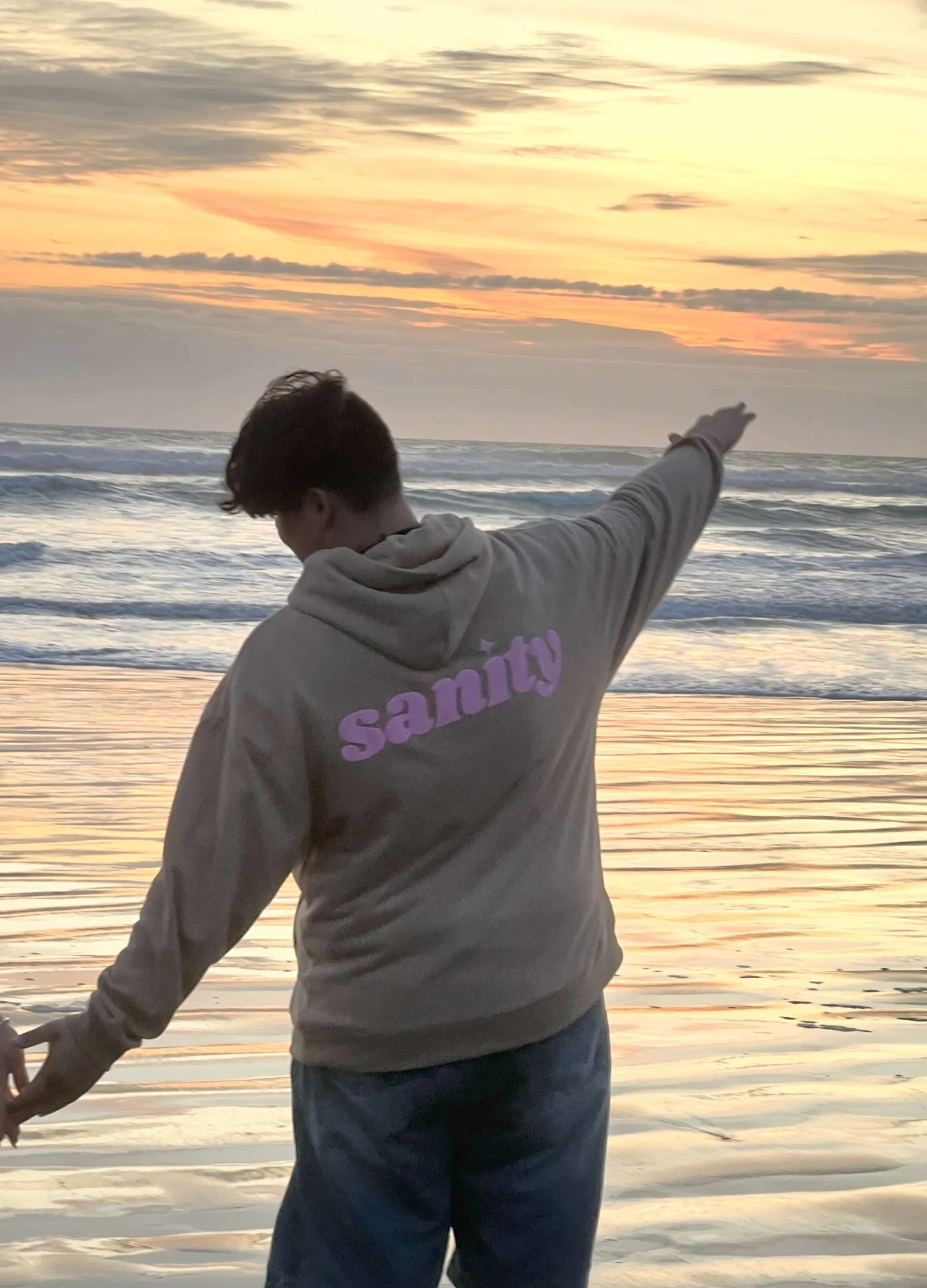 Person wearing Sanity chocolate hoodie with pink print by the ocean at sunset, showcasing comfy style and luxury.