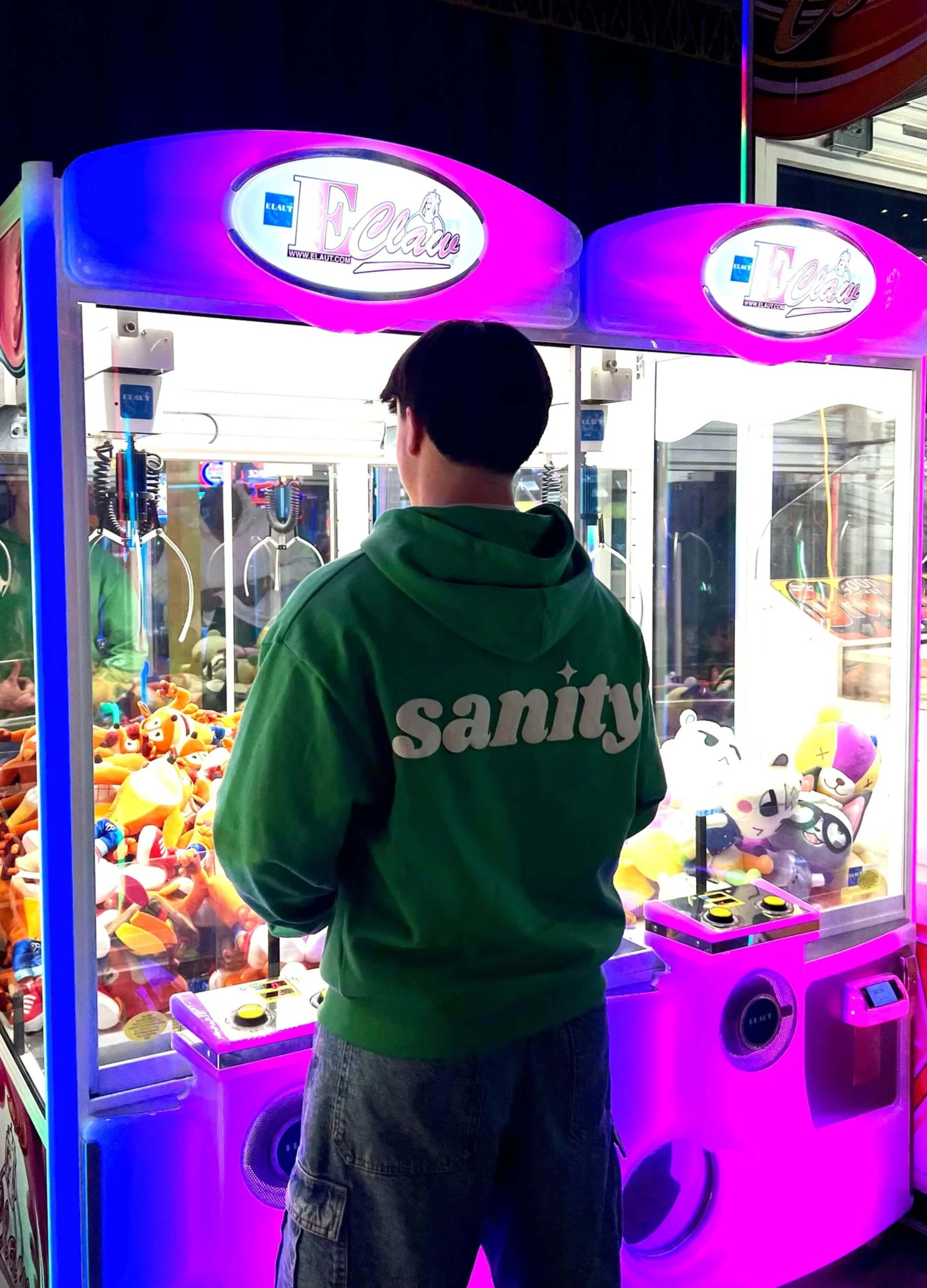 A person wearing a green Sanity zip-up hoodie stands by colorful arcade machines.