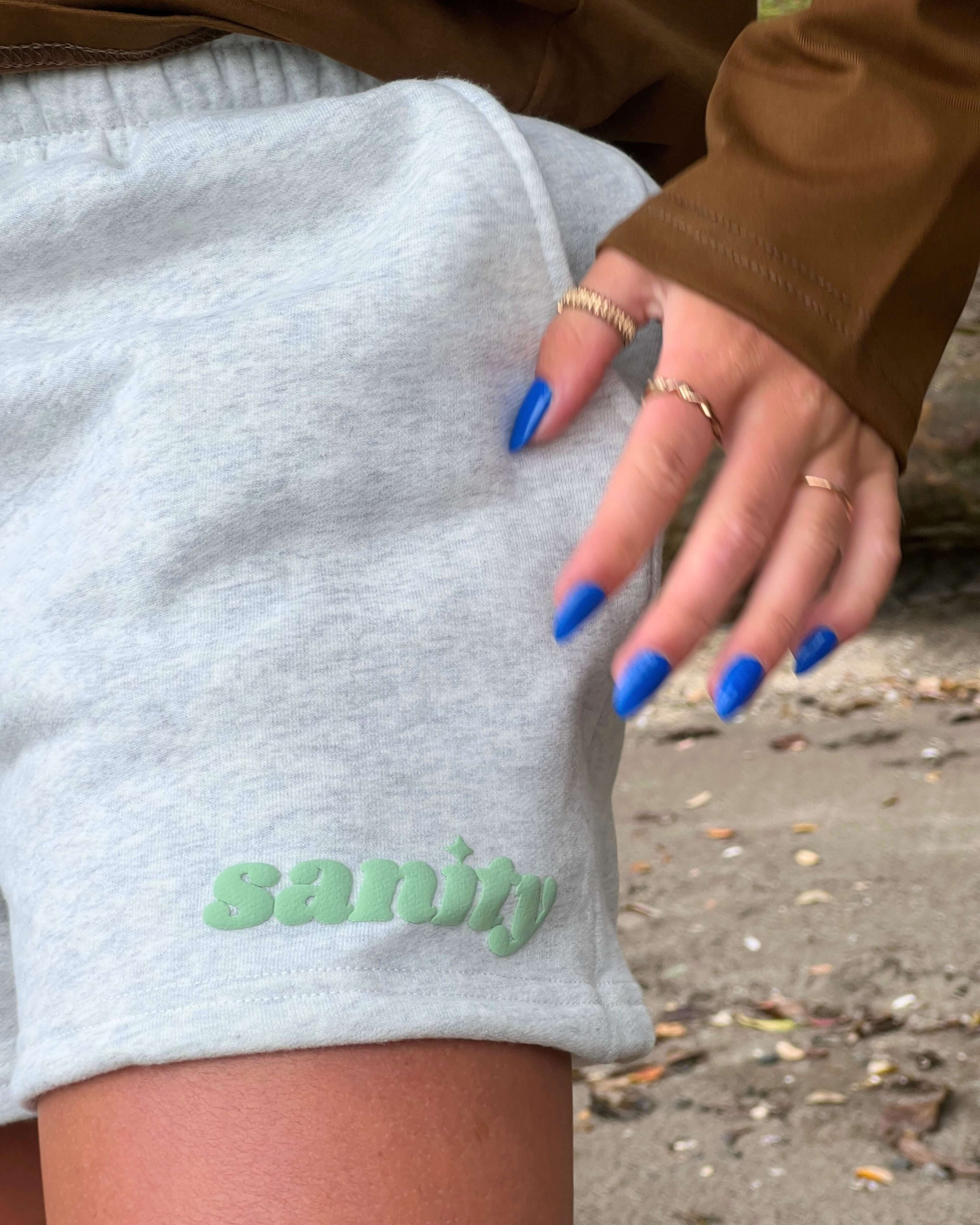 Close-up of a woman's hand on gray Sanity shorts, showcasing the logo and stylish nails against a natural background.