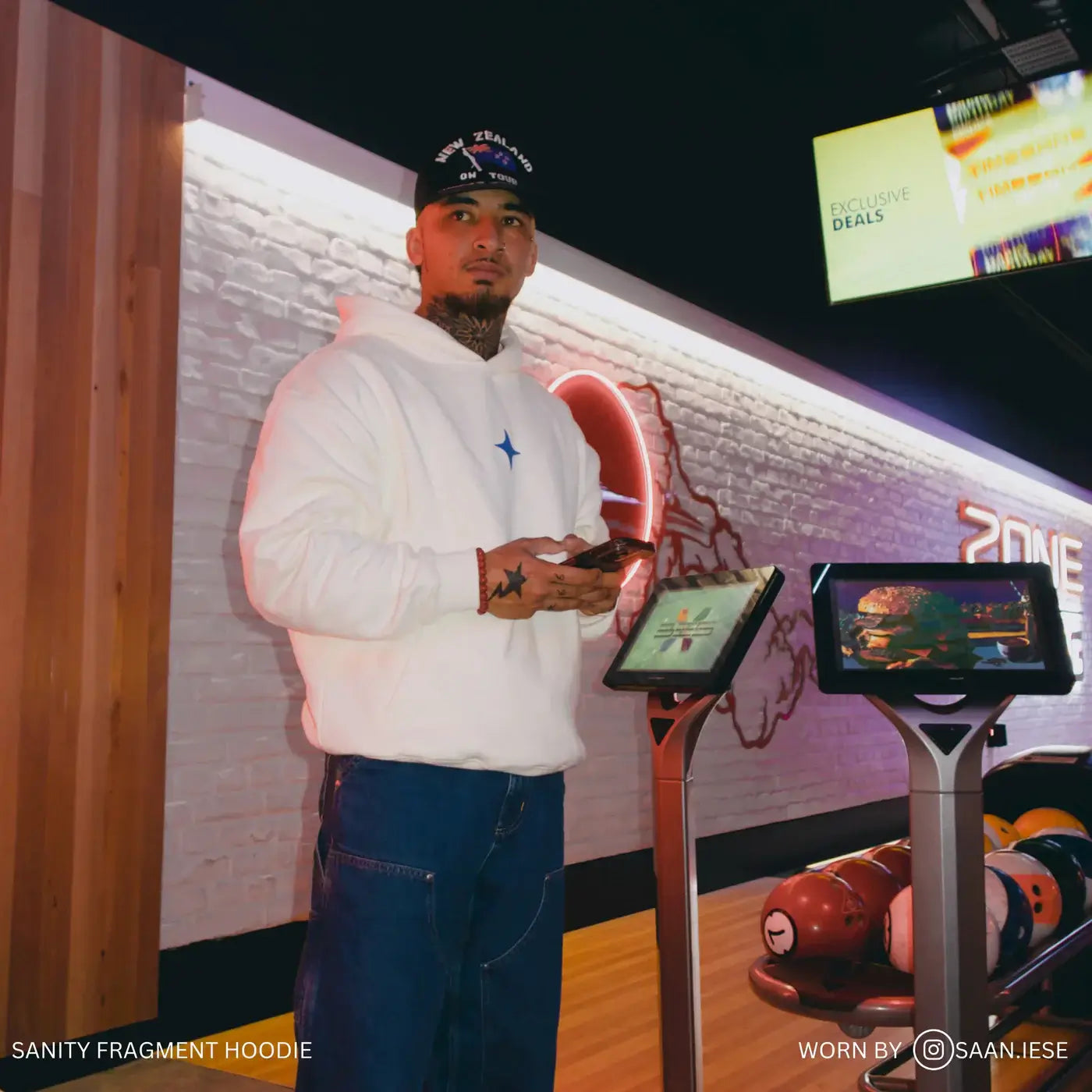 Man wearing Sanity Fragment Hoodie in cream with blue print, standing in a bowling alley setting.
