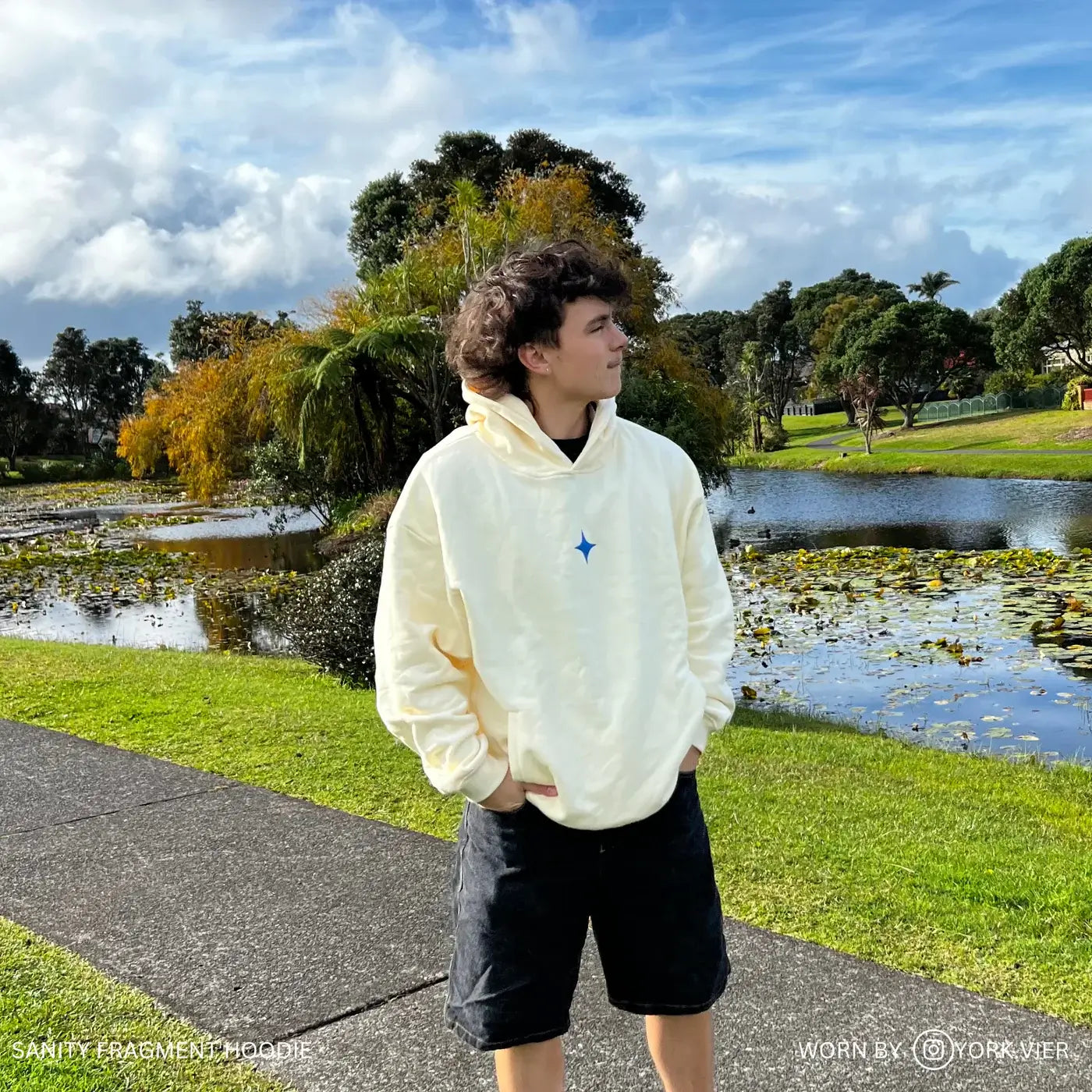 Unisex Sanity Fragment Hoodie in cream with blue print, displayed outdoors by a young man near a tranquil pond.