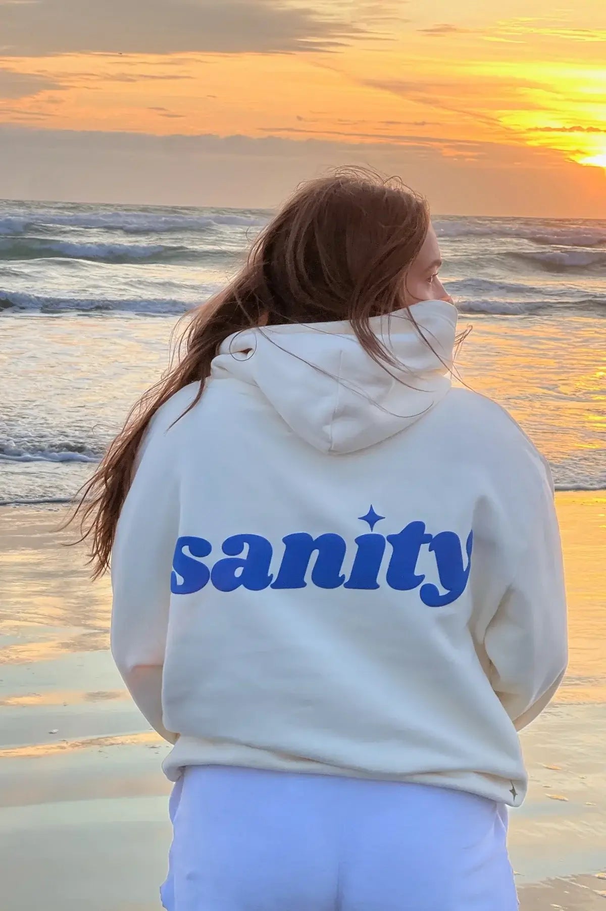 Back view of a woman wearing a cream Sanity hoodie with blue print, standing on the beach at sunset.