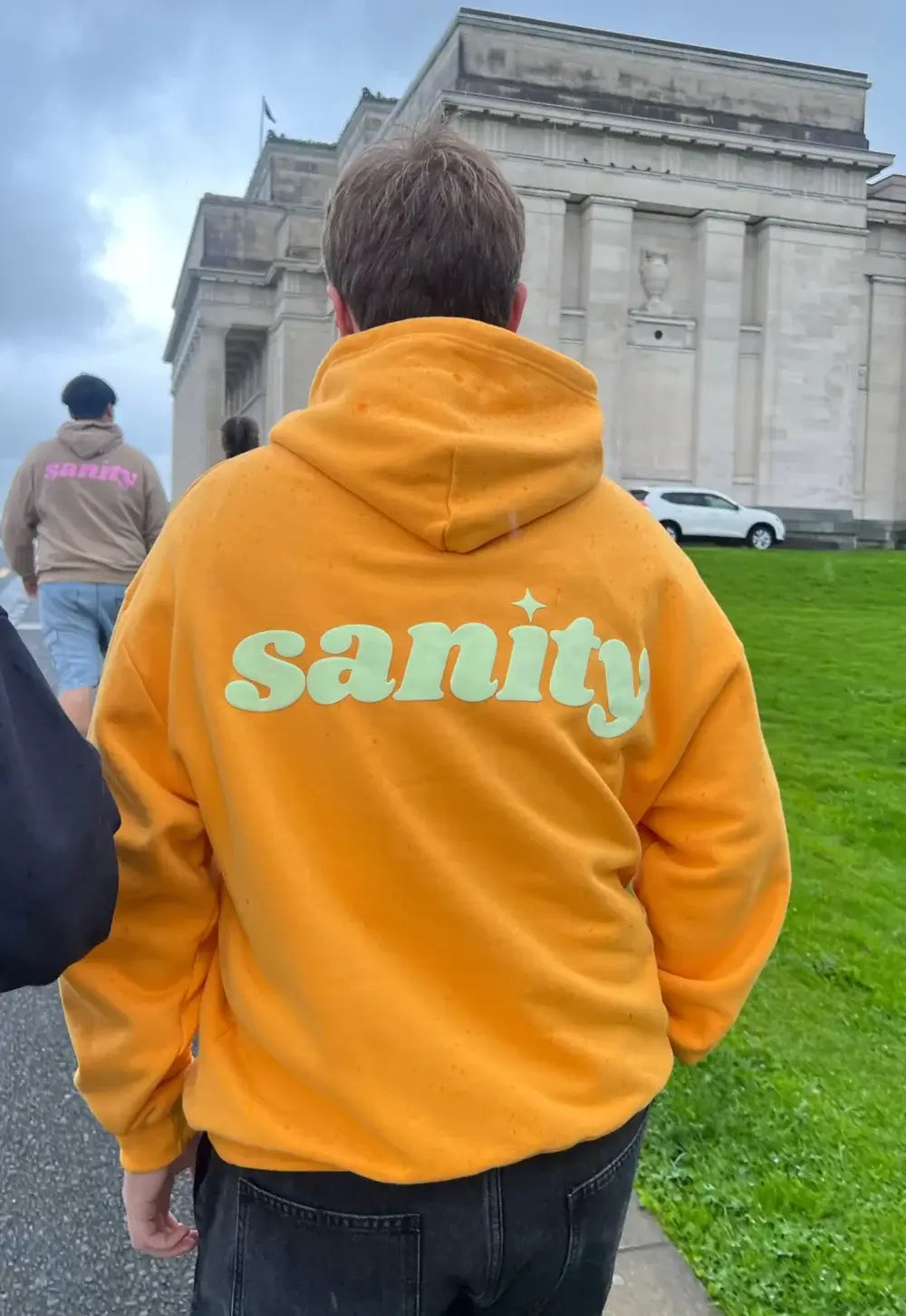 Person wearing an orange Sanity hoodie with mint logo, walking outdoors in a casual setting.