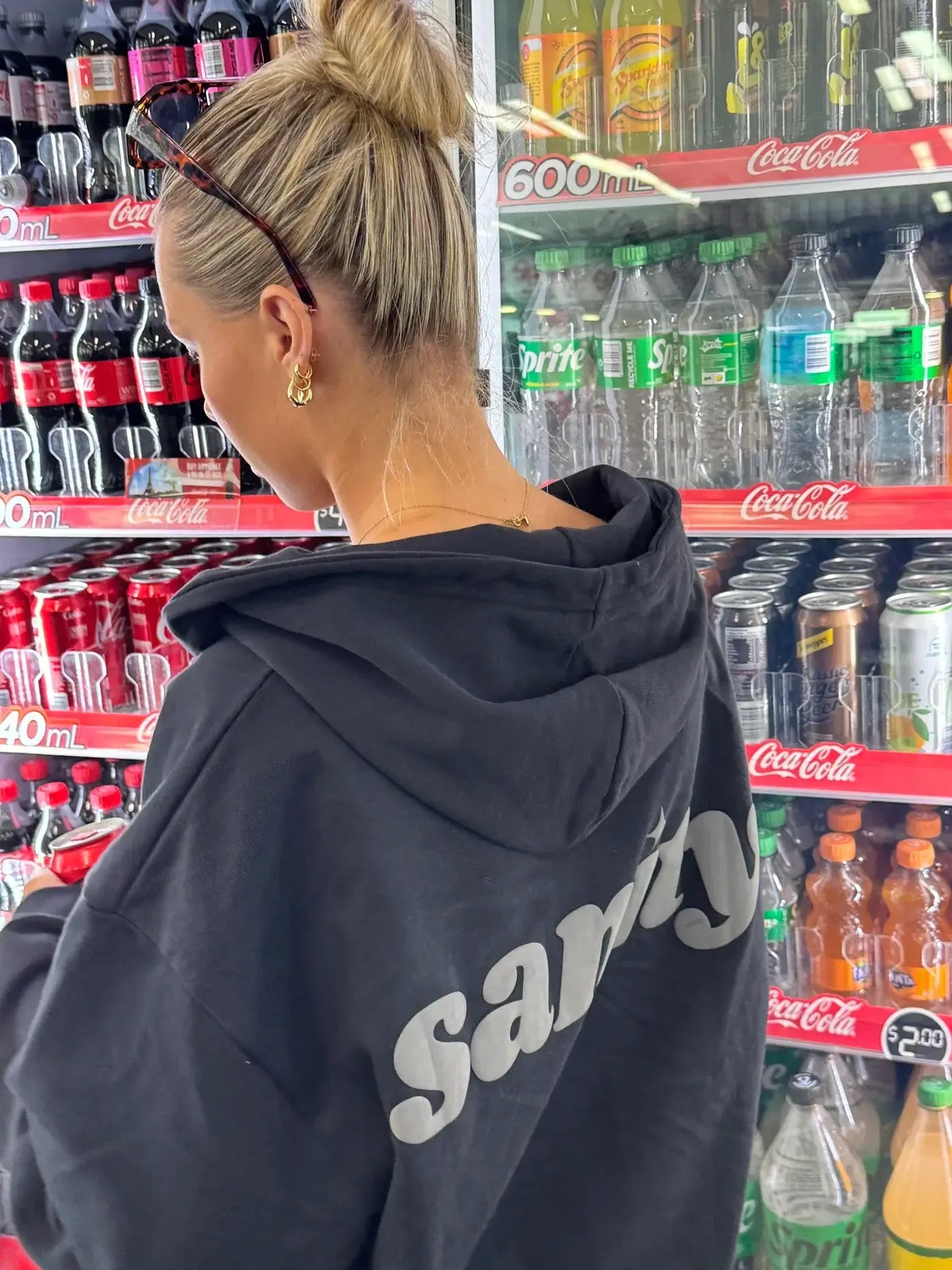Woman wearing Sanity midnight zip-up hoodie with grey puff print, standing in front of a beverage fridge.