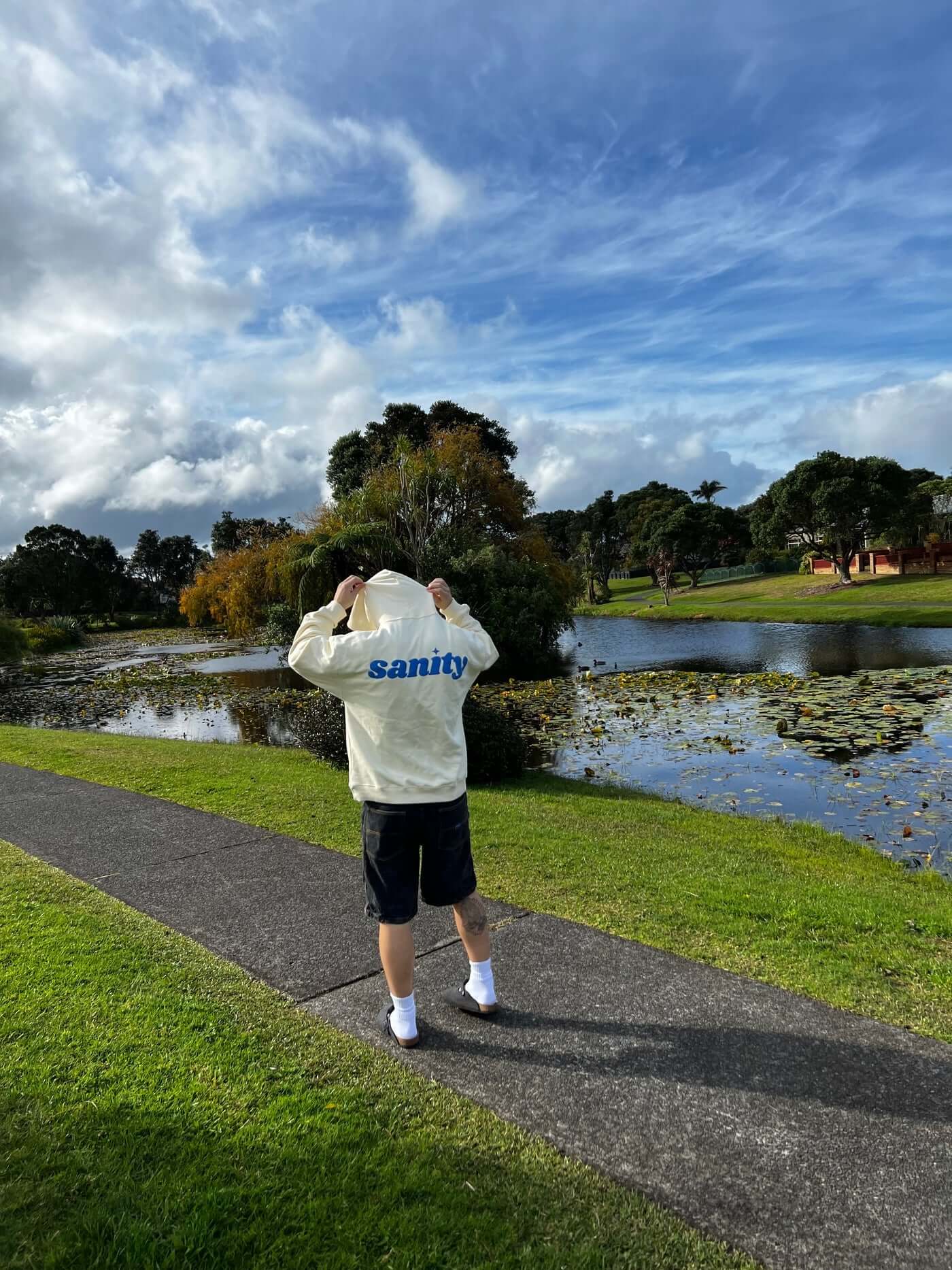 Person wearing a cream hoodie with blue print and 'sanity' embroidery by a scenic lake.