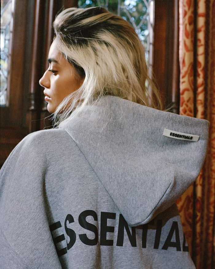 Woman wearing a gray hoodie with 'ESSENTIALS' tag, showcasing modern streetwear style against elegant background.